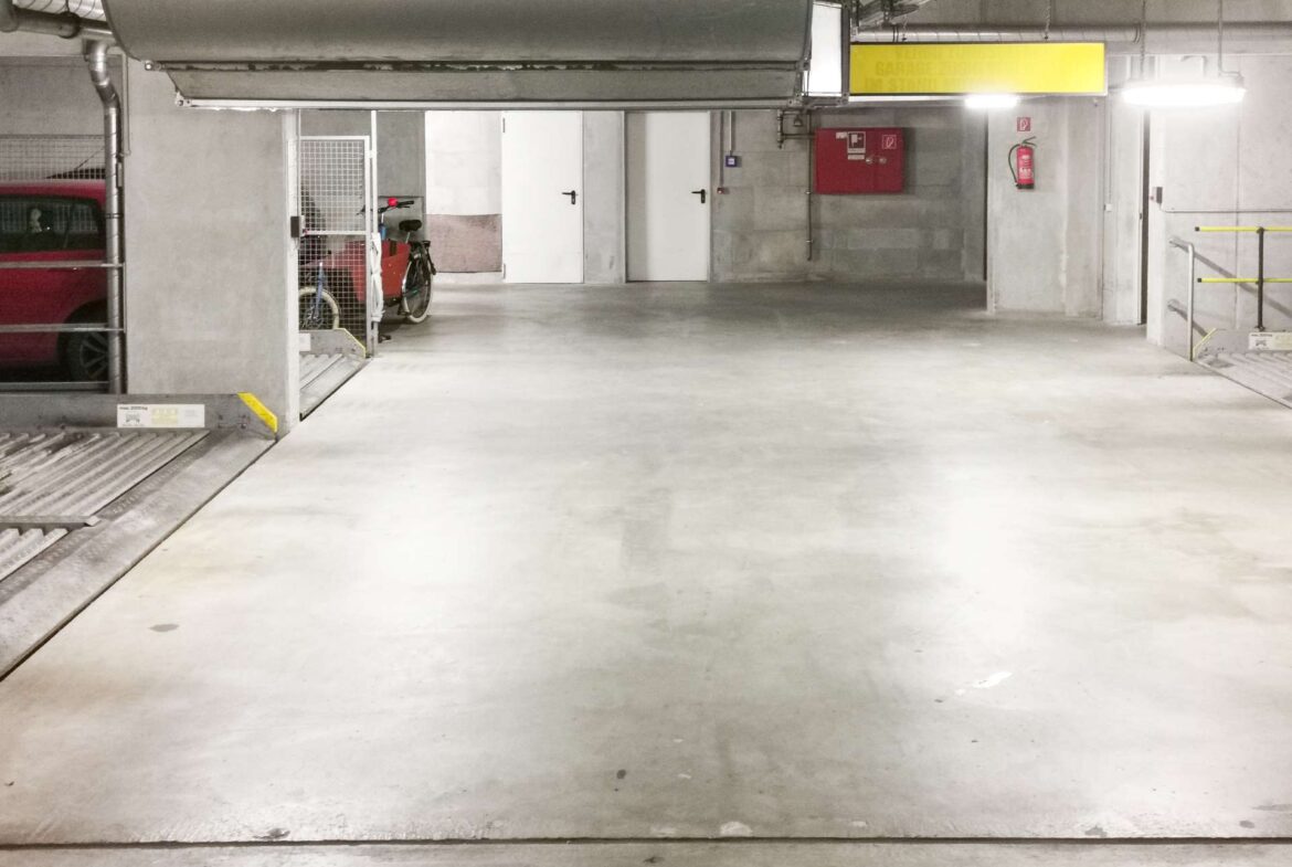 Underground parking area with a red van on the left and a bicycle behind a mesh gate, three white doors along the back wall, and a yellow sign overhead.