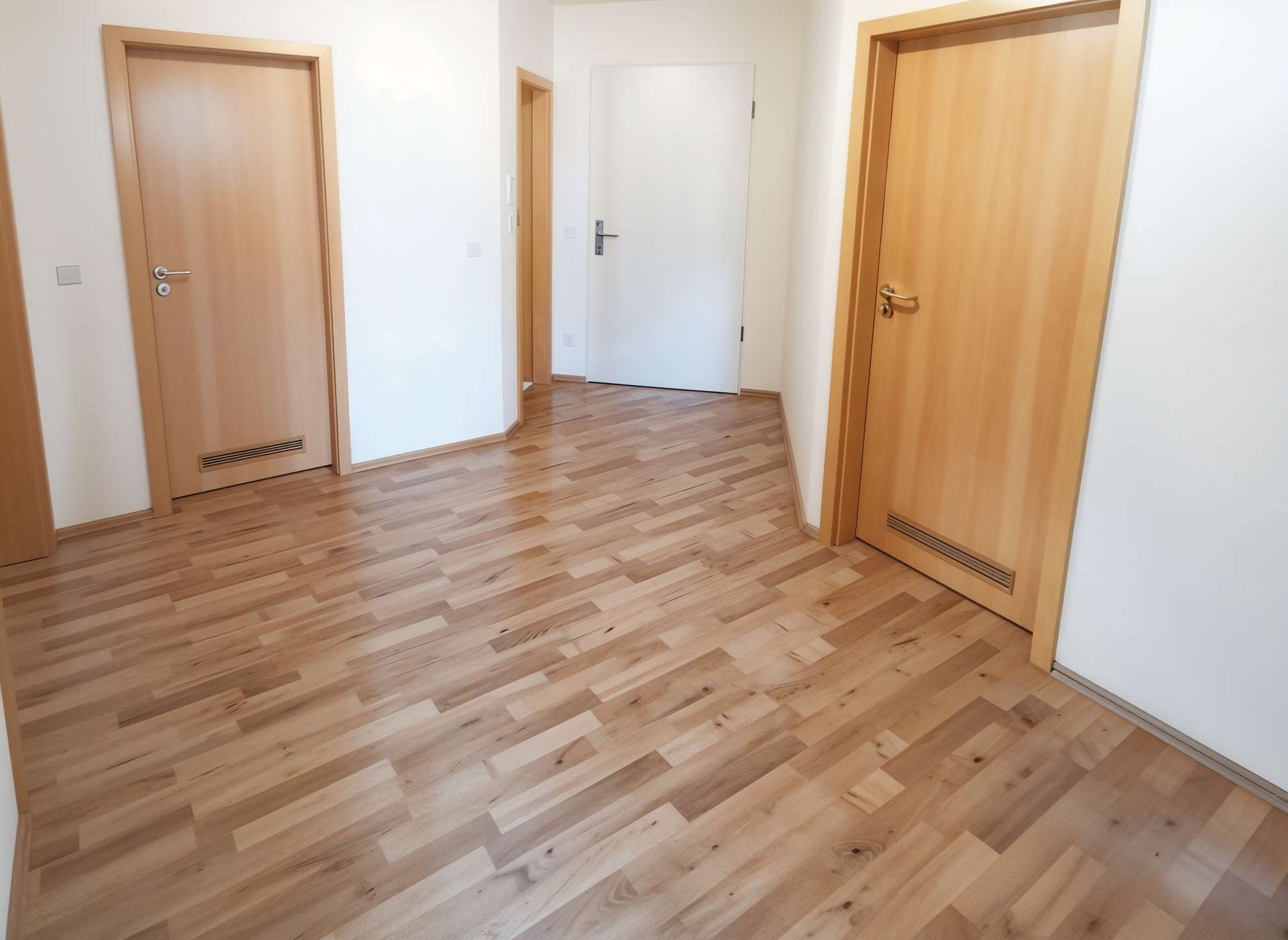Bright hallway with light wood flooring and three wooden doors along the walls, white walls surrounding.