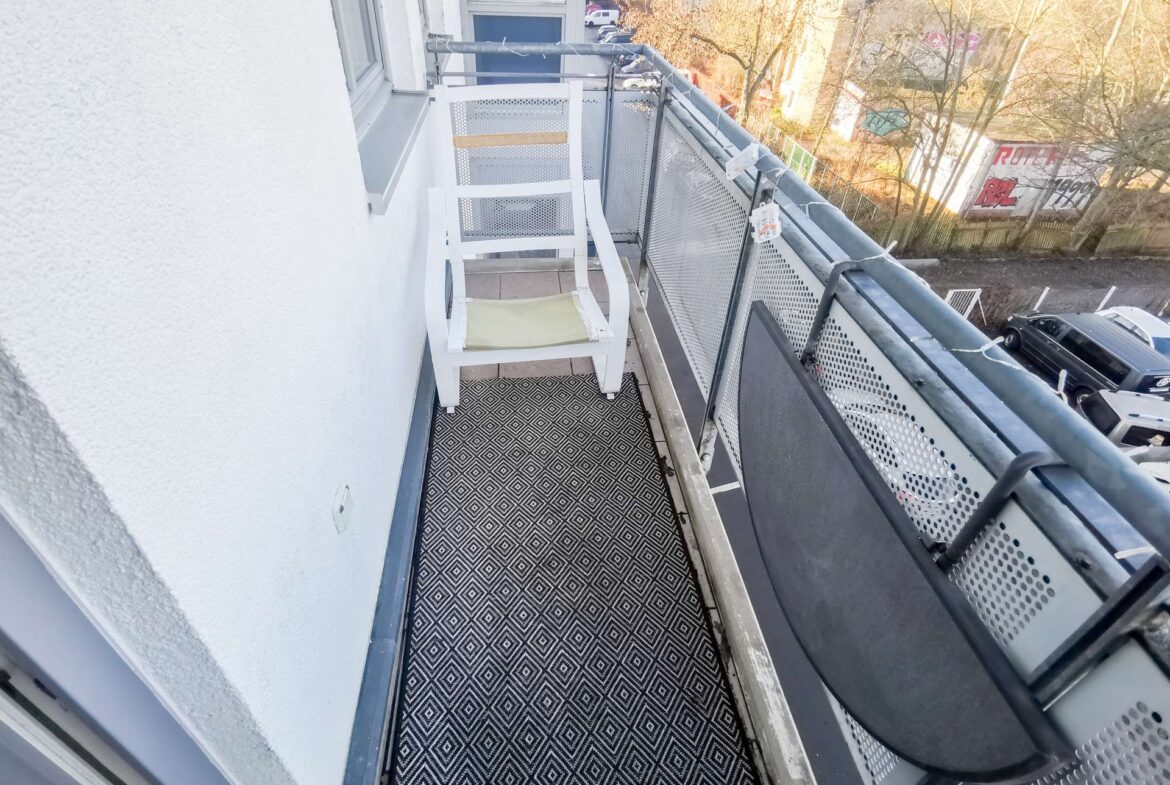 Narrow apartment balcony with a white chair, geometric-patterned floor, and a metal railing overlooking a street.