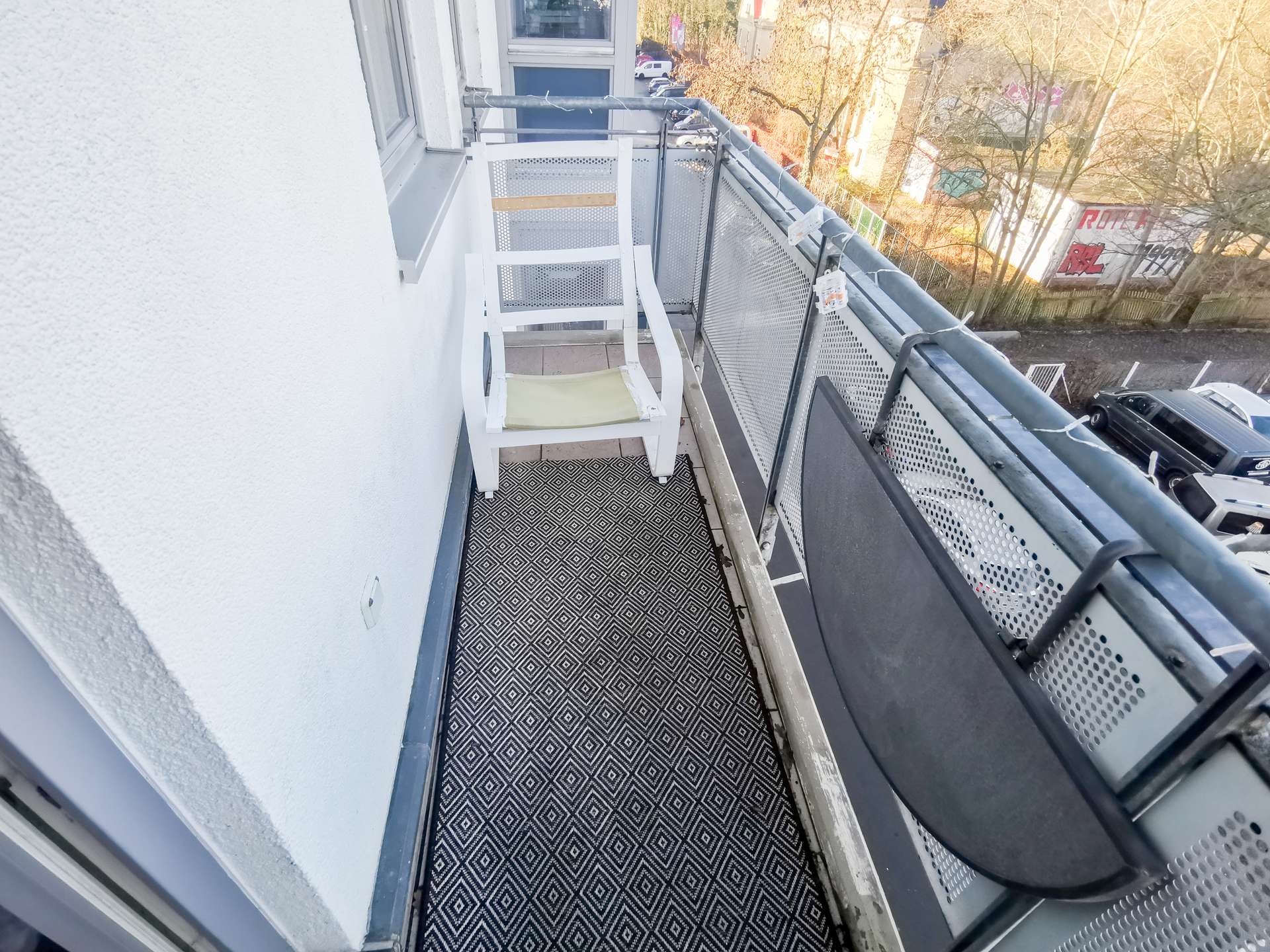 Narrow apartment balcony with a white chair, geometric-patterned floor, and a metal railing overlooking a street.