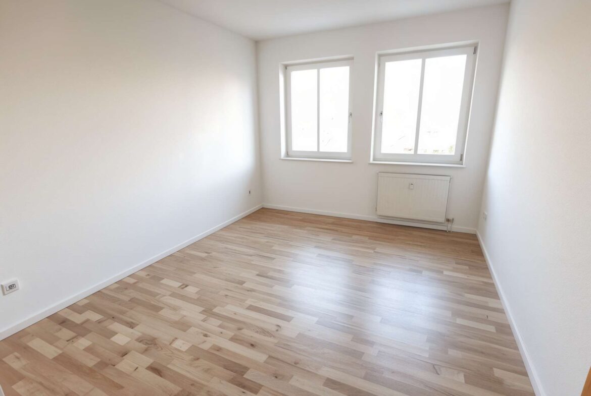 Bright empty room with white walls, light wood laminate floor, two large windows and a wall radiator beneath.