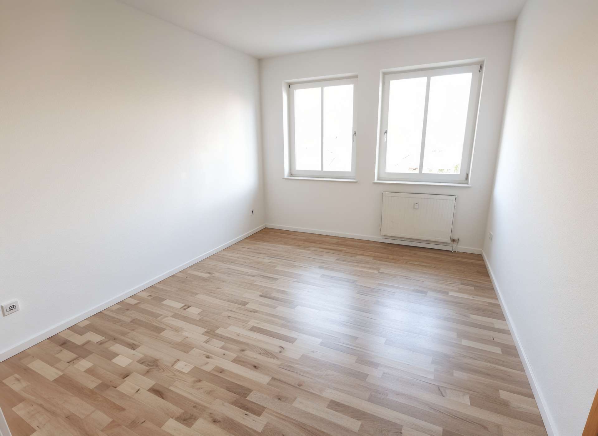 Bright empty room with white walls, light wood laminate floor, two large windows and a wall radiator beneath.