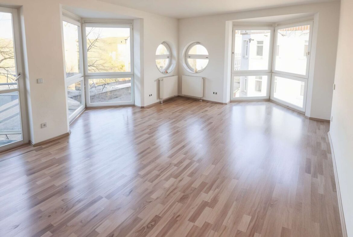 Bright empty living room with large bay windows and two round windows above radiators in white walls and light wood flooring