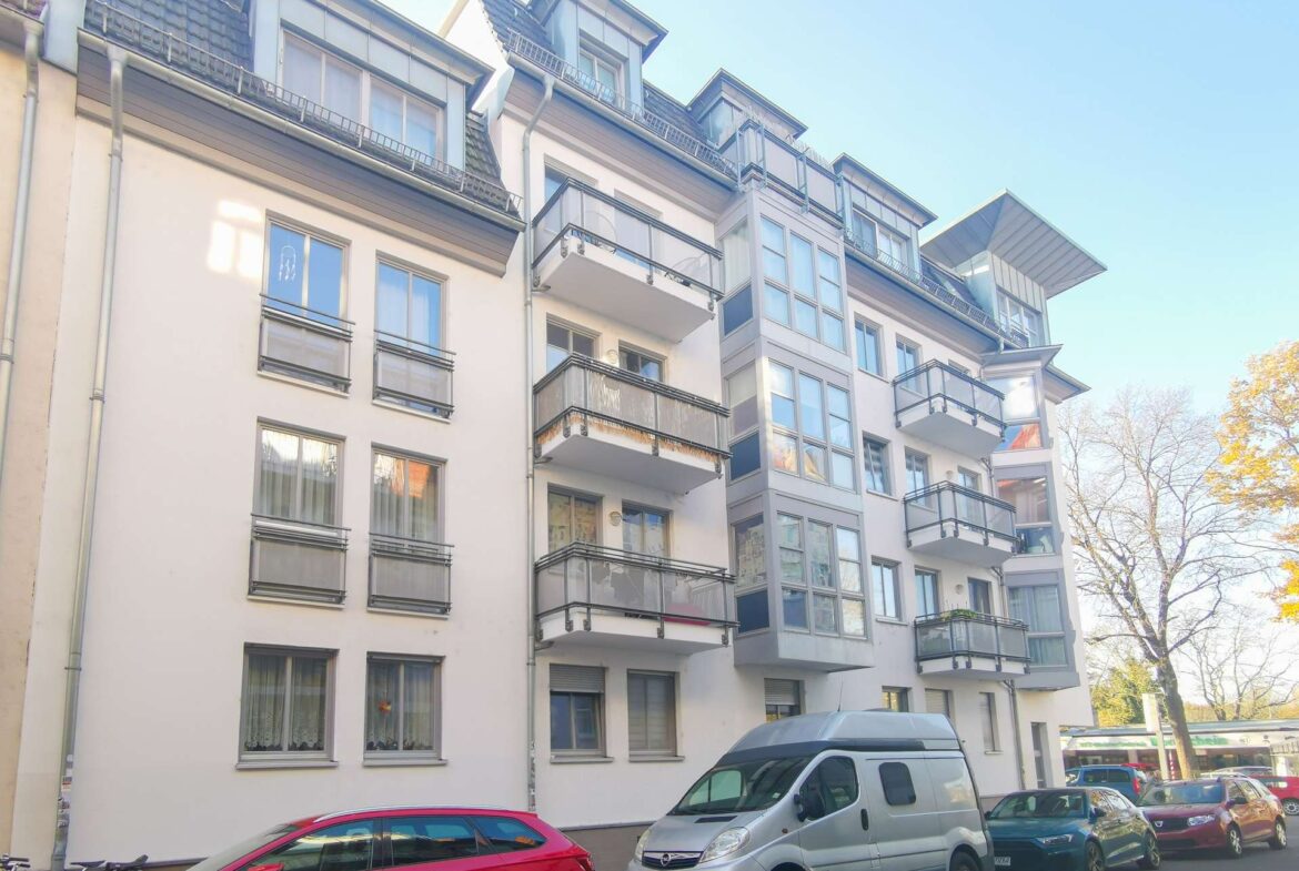 Modern white apartment building with glass-front balconies along a tree-lined street, cars parked outside on a sunny day.