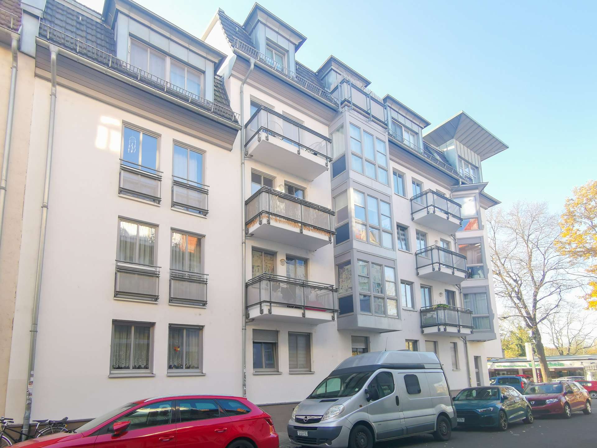 Modern white apartment building with glass-front balconies along a tree-lined street, cars parked outside on a sunny day.