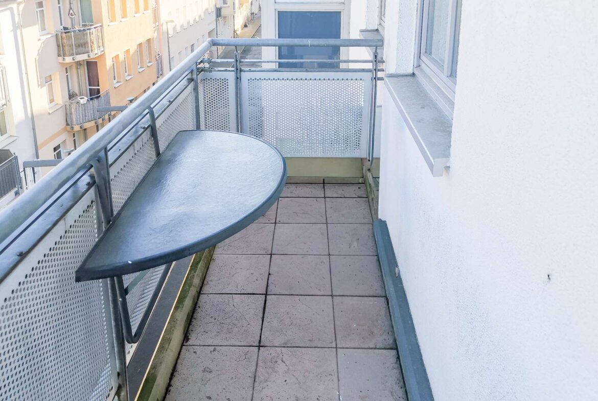 Balcony view with a round metal table and perforated metal railing, overlooking pastel apartment buildings across the street.