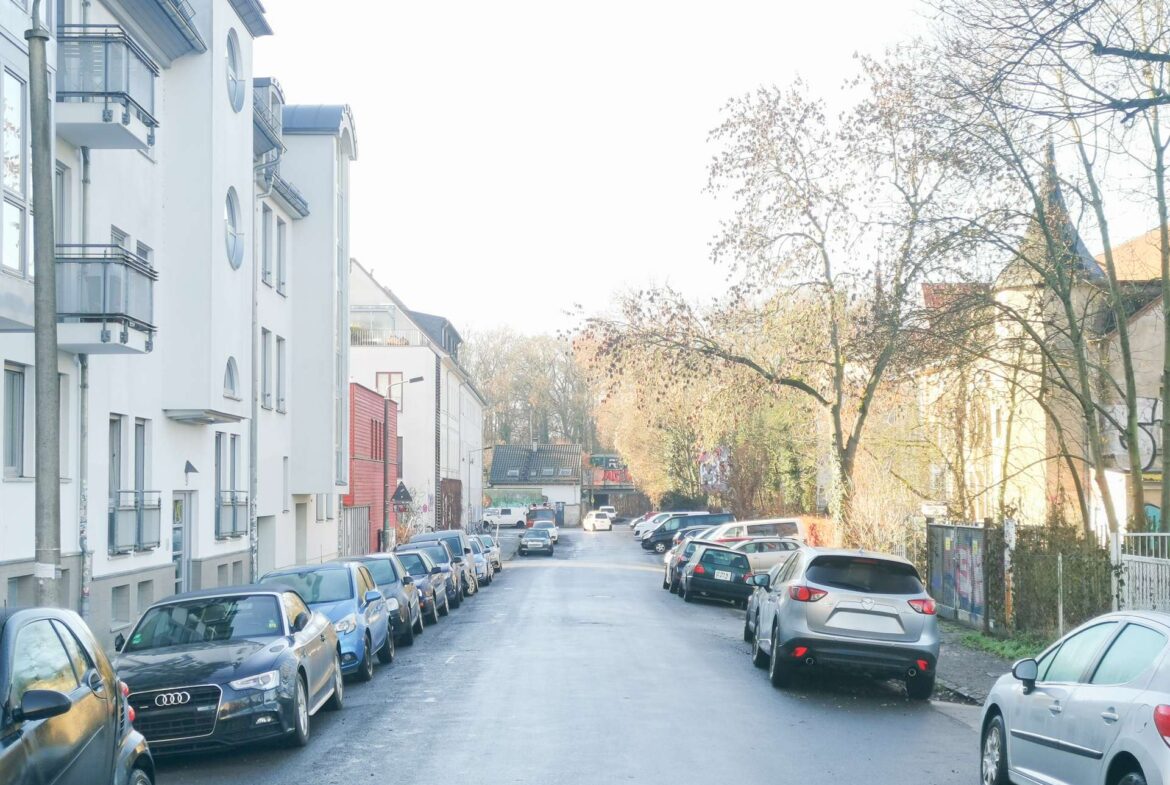 Residential street lined with parked cars on both sides, white apartment buildings to the left, and leafless trees on the right.