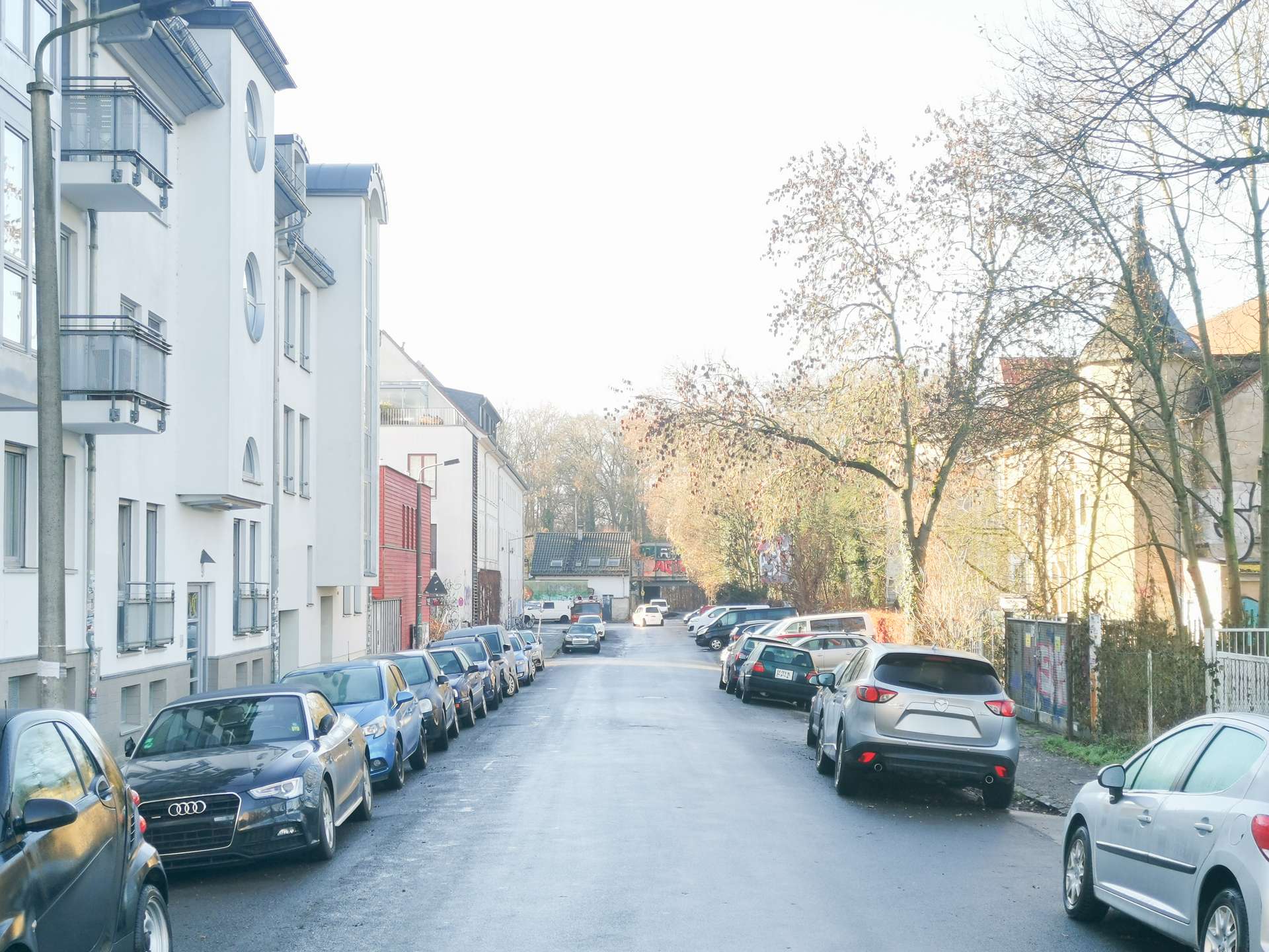 Residential street lined with parked cars on both sides, white apartment buildings to the left, and leafless trees on the right.