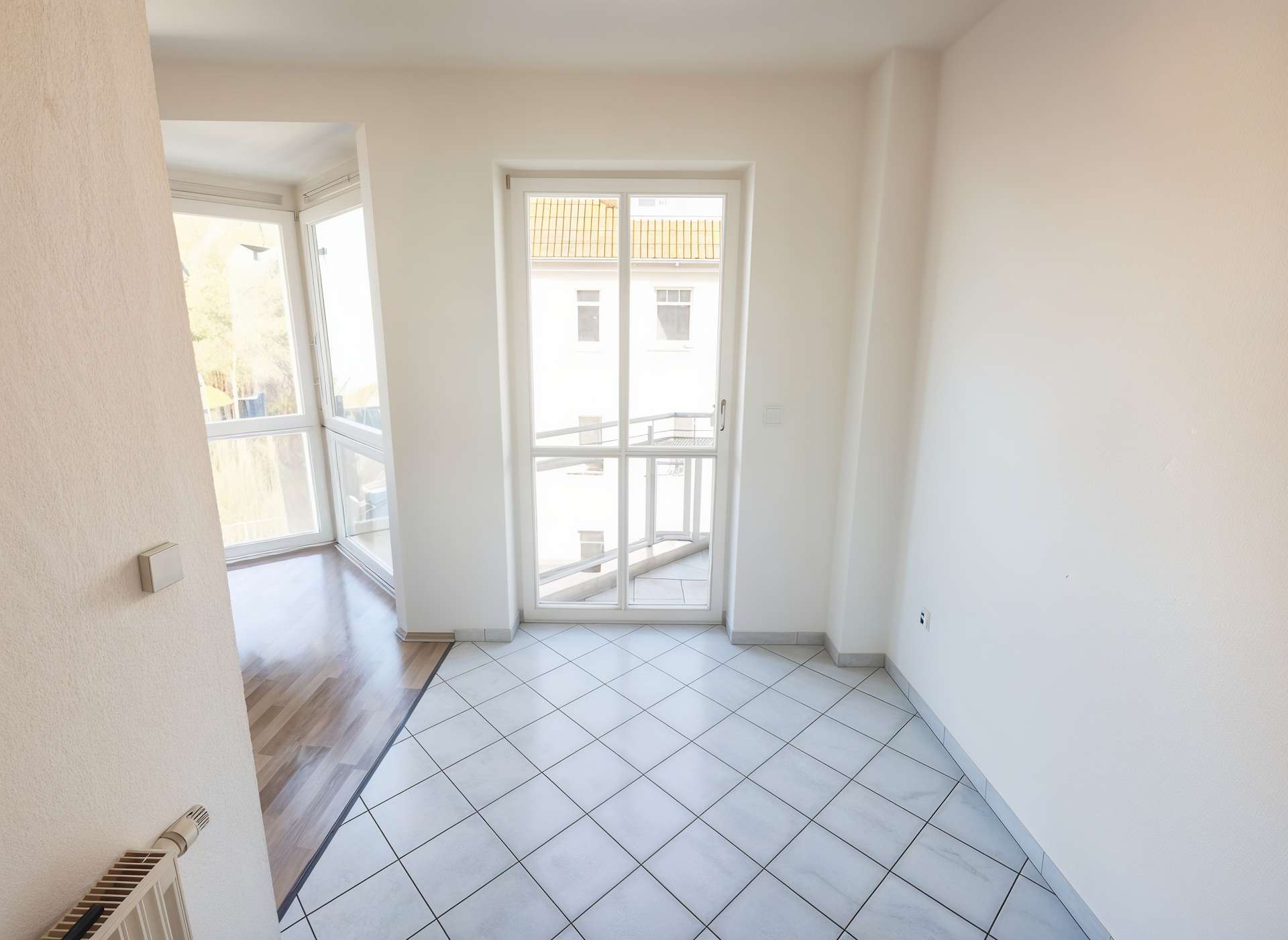 Bright corner room with large white-framed glass doors opening to a balcony, white walls and tiled floor.