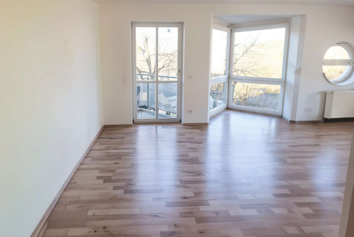 Bright empty living room with light wood floors, a balcony door and large corner window showing trees outside; round window with radiator on the right.