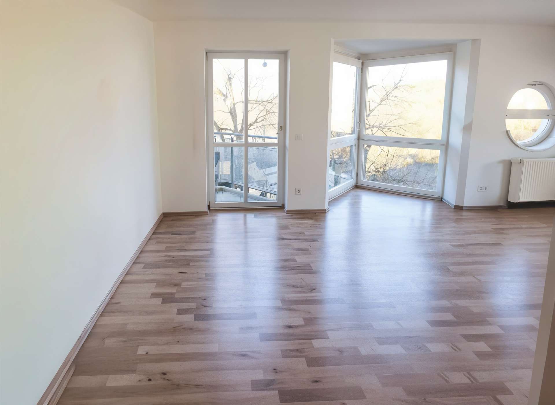 Bright empty living room with light wood floors, a balcony door and large corner window showing trees outside; round window with radiator on the right.