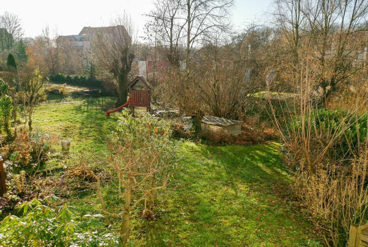 Backyard garden with a wooden playset and red slide, surrounded by bare trees and green grass, houses in the background on a sunny day.