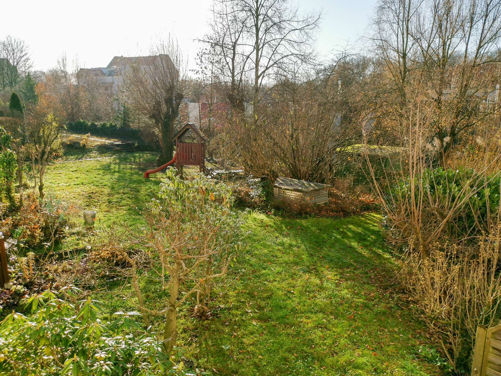 Backyard garden with a wooden playset and red slide, surrounded by bare trees and green grass, houses in the background on a sunny day.