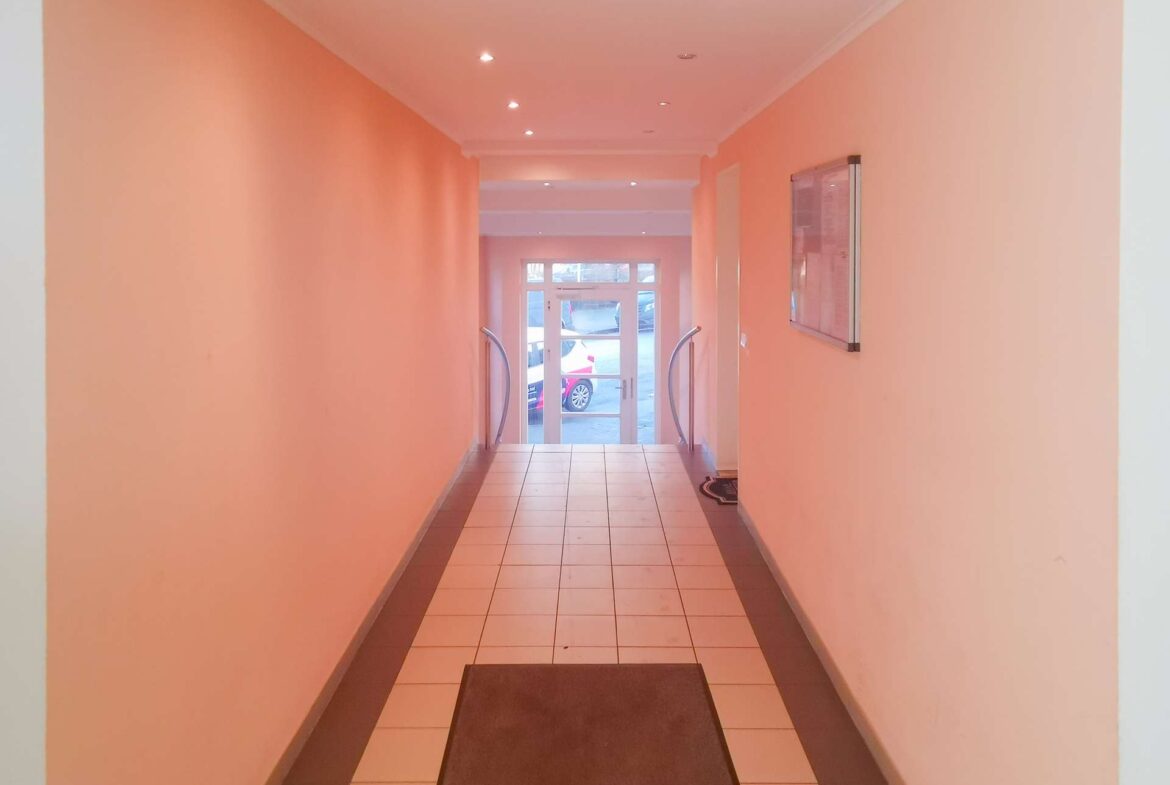 Narrow peach hallway with tiled floor and brown runners leading to a glass door at the far end, flanked by curved handrails on both sides.