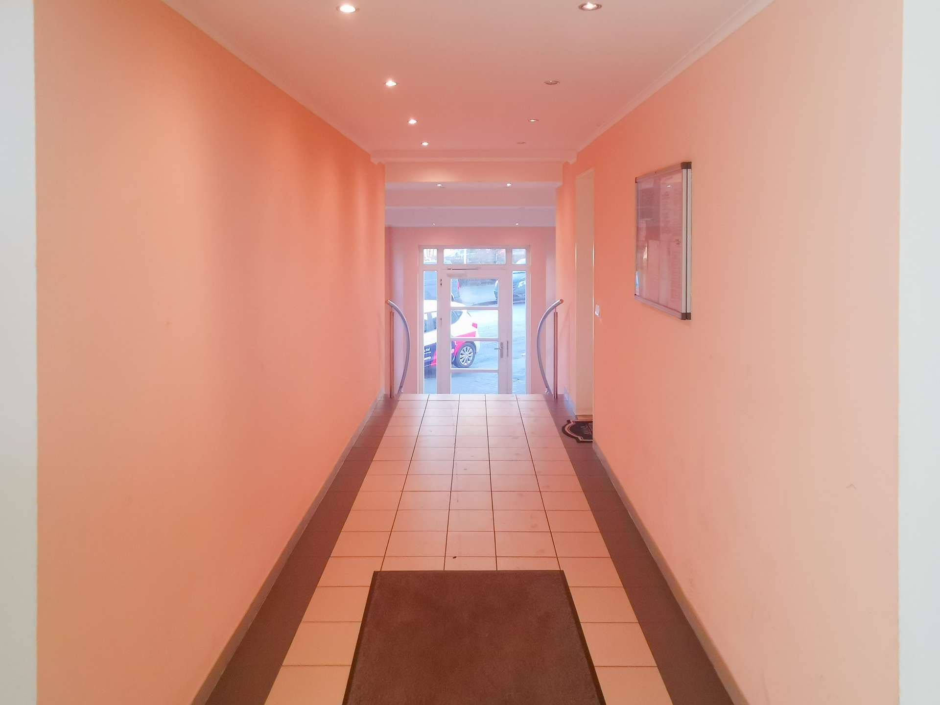 Narrow peach hallway with tiled floor and brown runners leading to a glass door at the far end, flanked by curved handrails on both sides.