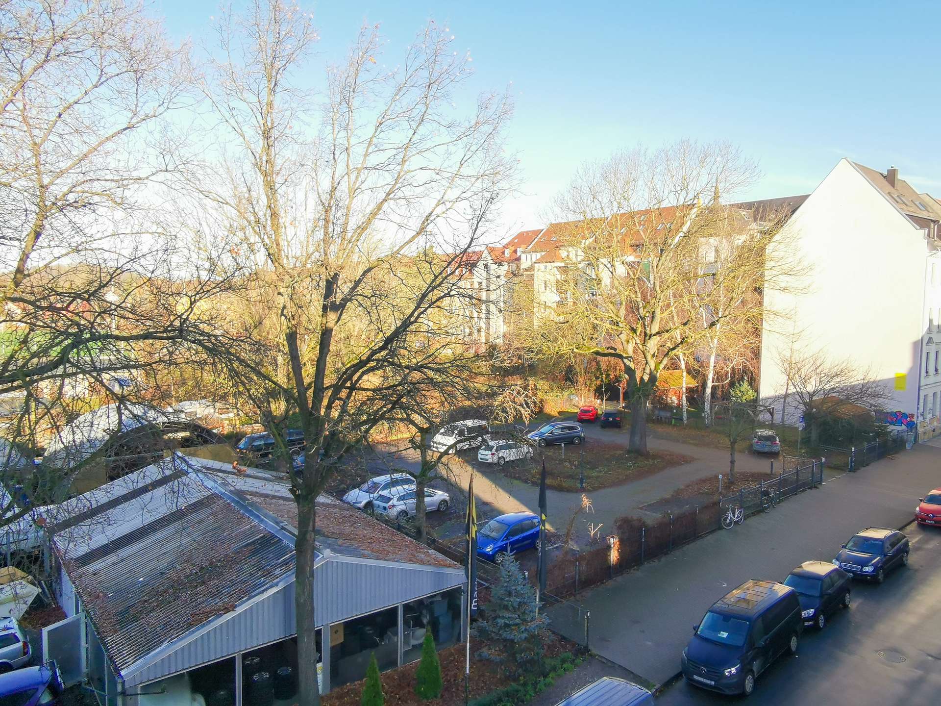 Residential street scene in winter: leafless trees lining a sunlit road with parked cars and row houses with red roofs in the background.