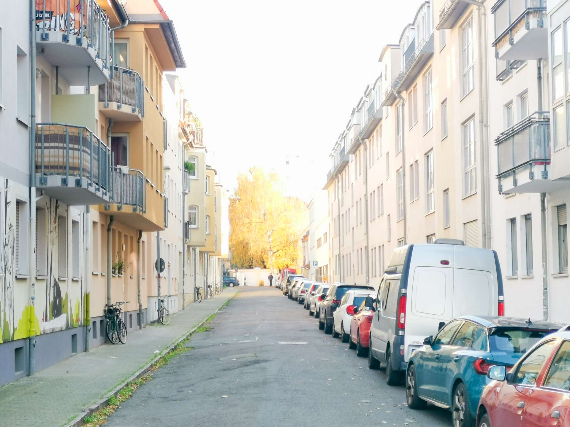 Urban residential street lined with pastel apartment buildings, with cars parked along both sidewalks and a white van in the foreground.