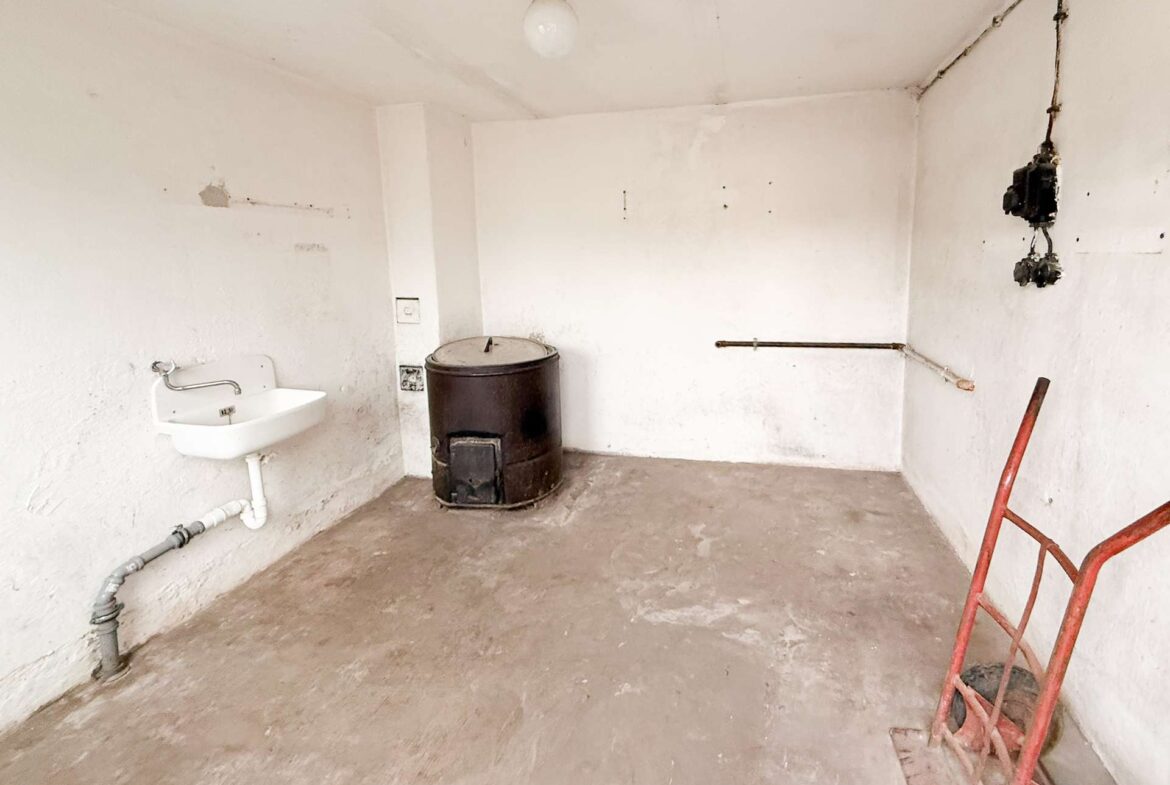 Empty utility room with a white wall-mounted sink on the left, a metal barrel stove in the center, and a red cart on the right.