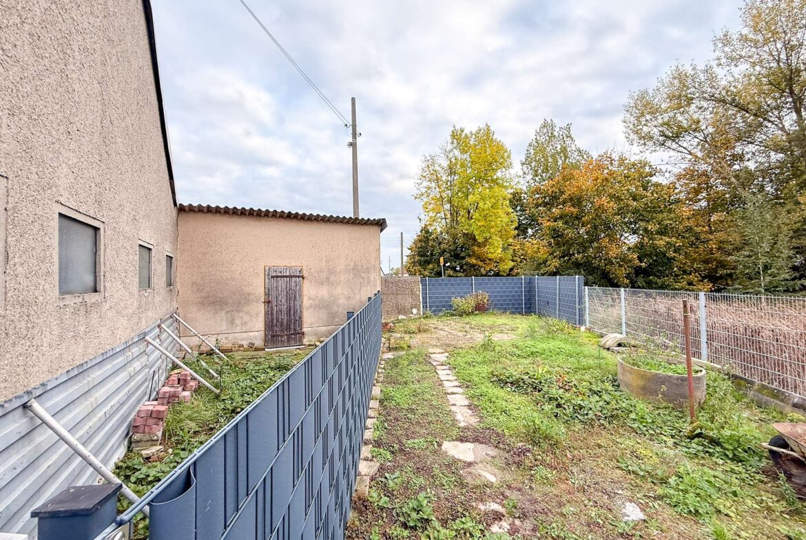 Beige stucco building on the left with small windows, a blue metal fence along a rocky yard, and an overgrown path toward a wooden door. Overhead cloudy sky and autumn trees in the background.