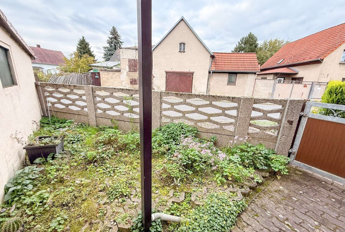 Backyard with overgrown plants and a stone-patterned wall, fence, and red-roofed houses in the background.