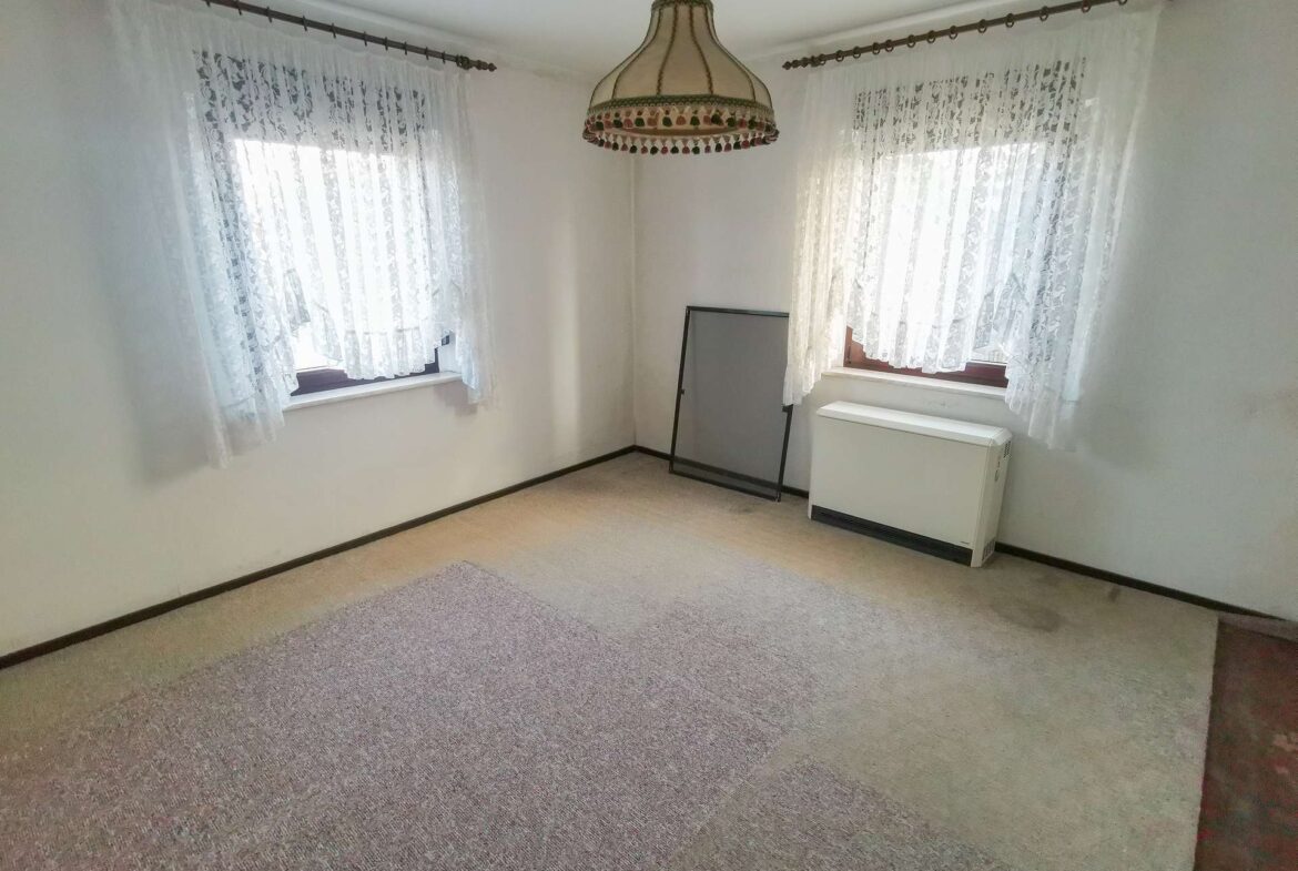 Empty beige carpeted room with two windows covered by lace curtains, and a vintage pendant light hanging from the ceiling.