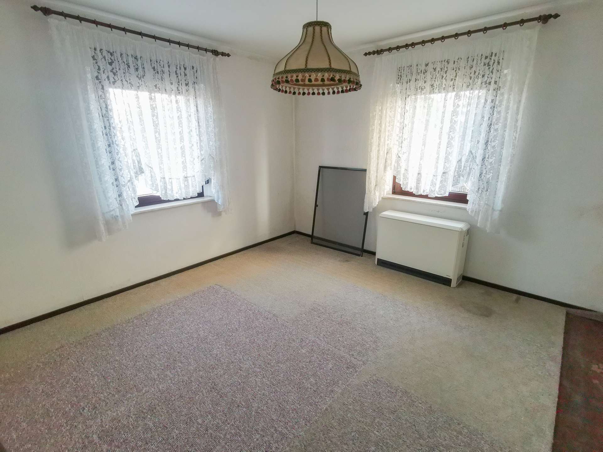 Empty beige carpeted room with two windows covered by lace curtains, and a vintage pendant light hanging from the ceiling.