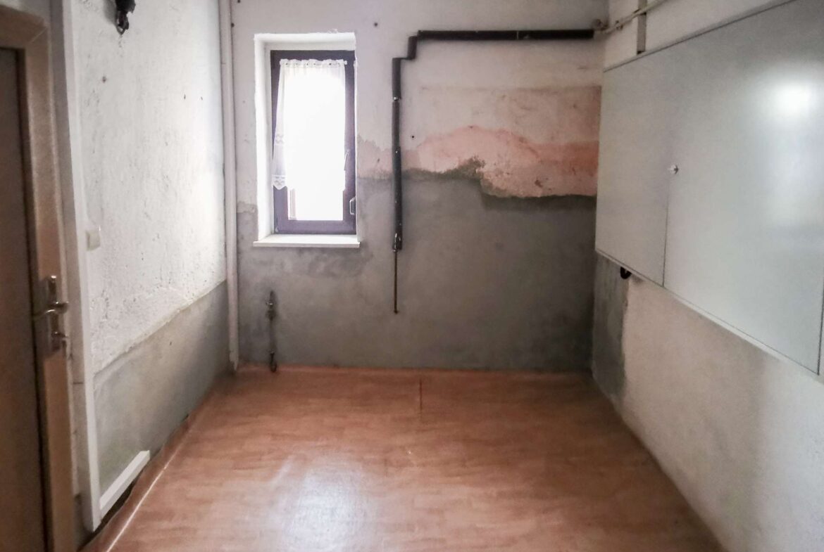 Narrow utility room with peeling plaster, a small window with lace curtain, and exposed pipes along a pale wall. A concrete half-wall and weathered surfaces dominate the space, with a wooden floor visible.