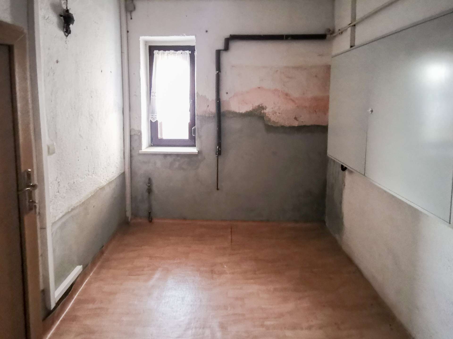 Narrow utility room with peeling plaster, a small window with lace curtain, and exposed pipes along a pale wall. A concrete half-wall and weathered surfaces dominate the space, with a wooden floor visible.
