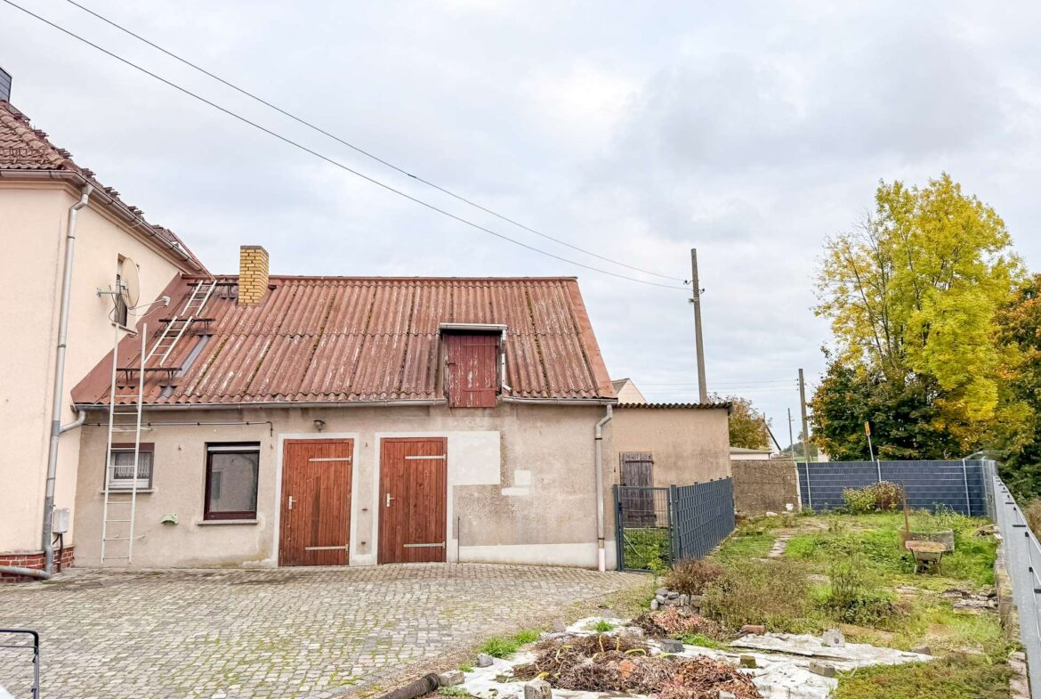 Beige building with a red tile roof and a ladder leaning on the wall; cobblestone surface in the foreground and a small satellite dish on the wall.
