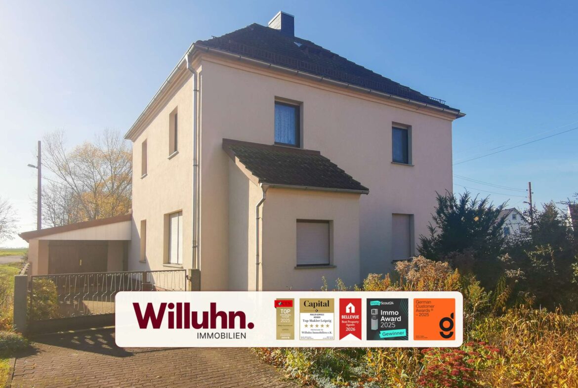 Two-story beige house under a clear blue sky with a large real estate sign in the front yard reading Willuhn Immobilien and award badges nearby.