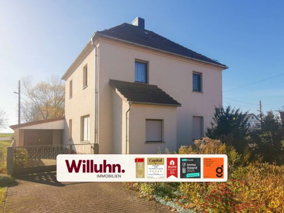 Two-story beige house under a clear blue sky with a large real estate sign in the front yard reading Willuhn Immobilien and award badges nearby.