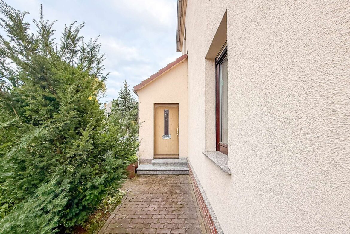 Entrance to a beige stucco house with a wooden door along a brick-paved walkway and a tall evergreen shrub on the left.
