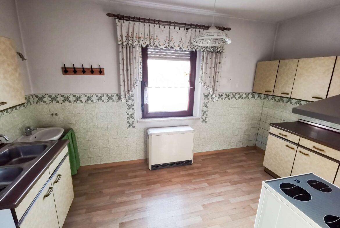 Old-fashioned kitchen with beige cabinets, tiled backsplash, a window with floral curtains, and a sink on the left.