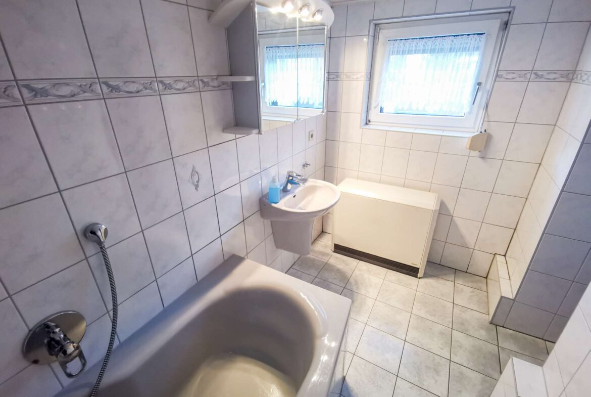 White tiled bathroom with a bathtub along the left, pedestal sink, washing machine, and a window with sheer blinds.