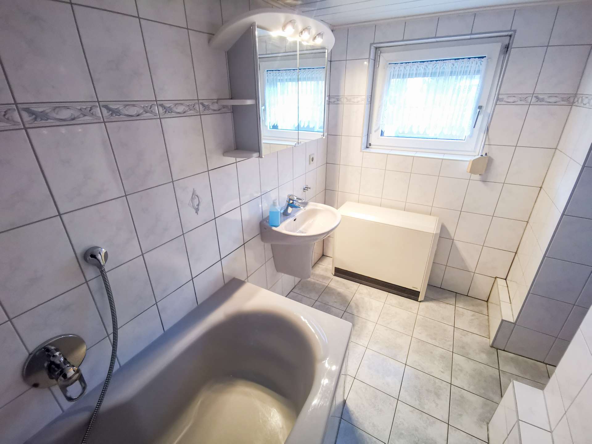 White tiled bathroom with a bathtub along the left, pedestal sink, washing machine, and a window with sheer blinds.