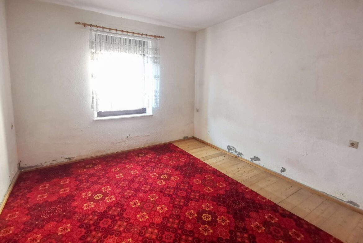 Empty white-walled room with a lace-curtained window and a red patterned carpet on a wooden floor