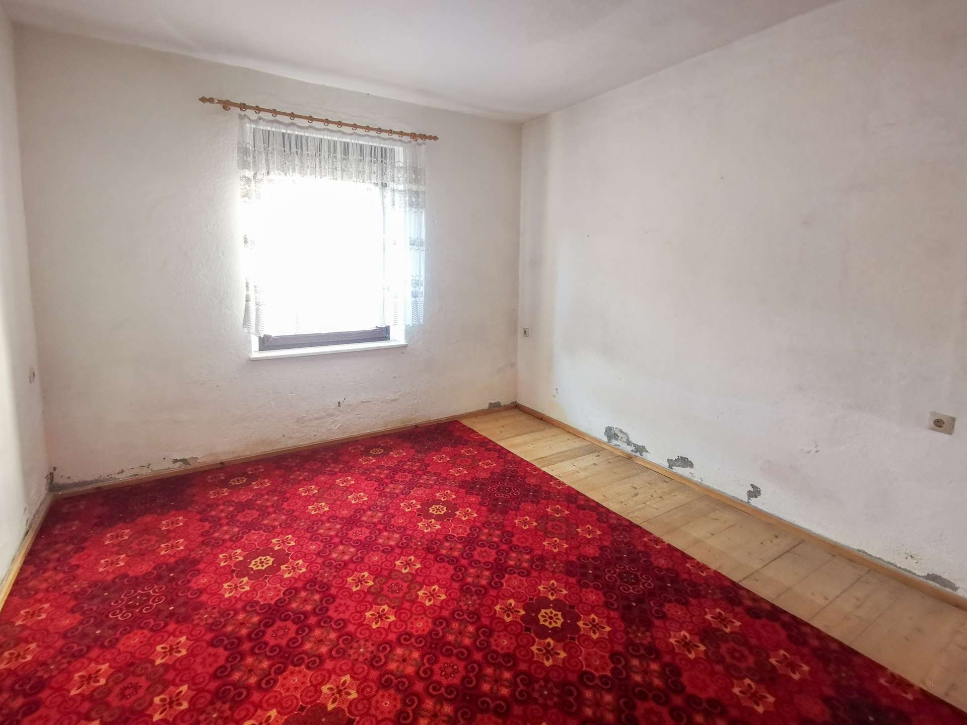 Empty white-walled room with a lace-curtained window and a red patterned carpet on a wooden floor