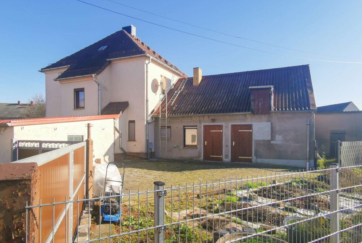 Front view of a two-story house with a sloped, dark roof, satellite dish, and a metal ladder leaning against the side, fenced yard in the foreground.
