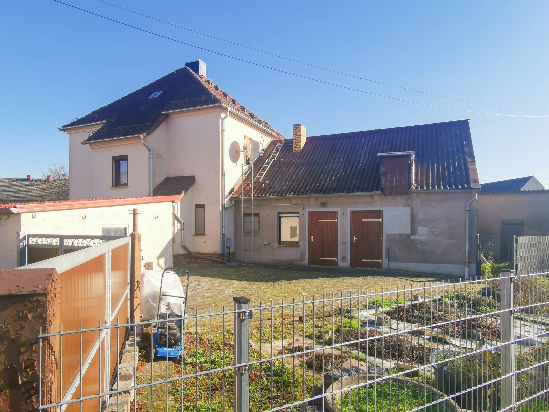 Front view of a two-story house with a sloped, dark roof, satellite dish, and a metal ladder leaning against the side, fenced yard in the foreground.