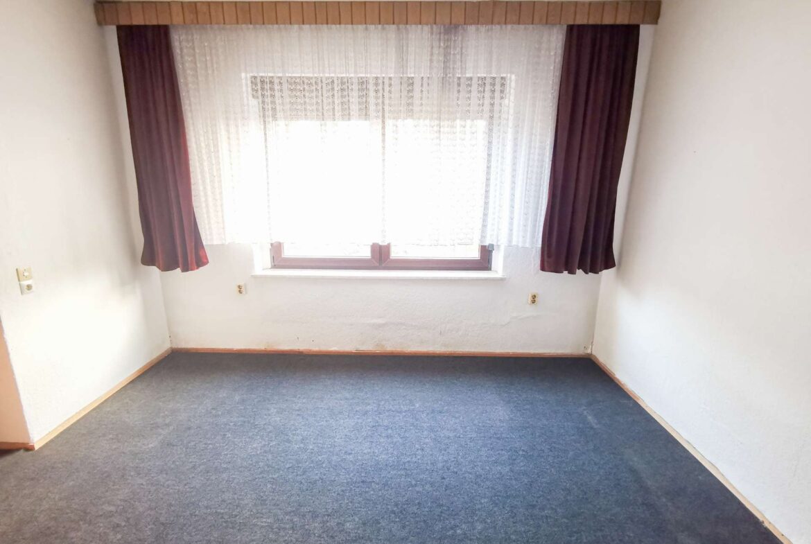 Small empty room with a large window covered by white lace and maroon curtains, blue carpet, white walls, and baseboard trim.