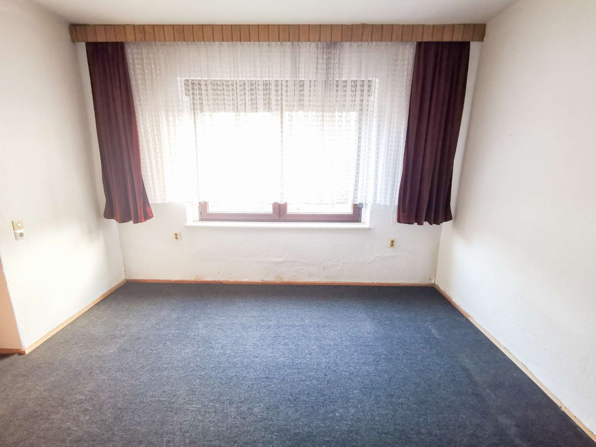 Small empty room with a large window covered by white lace and maroon curtains, blue carpet, white walls, and baseboard trim.