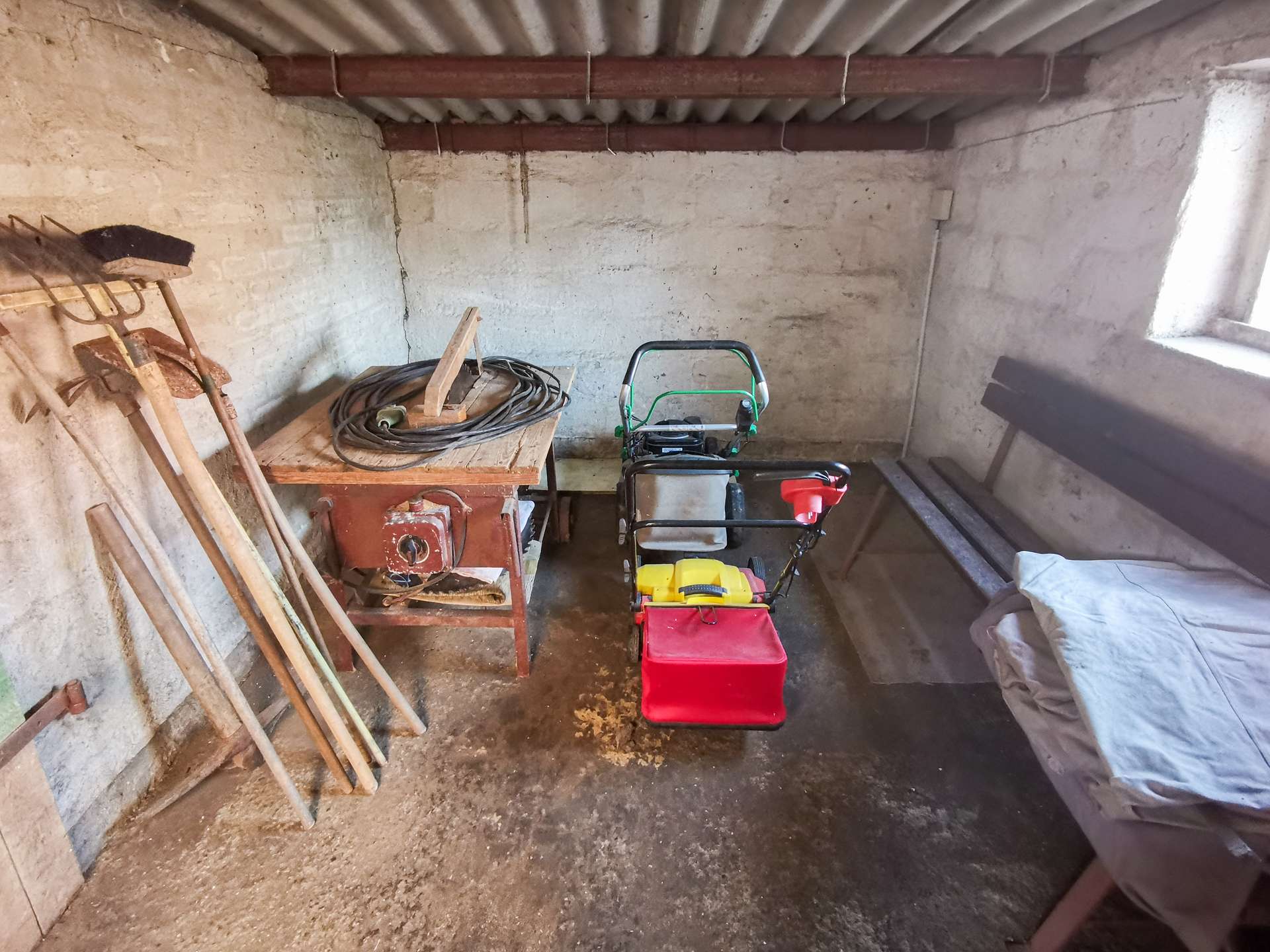 Dusty, cluttered workroom or shed with concrete walls and a corrugated roof; left wall has wooden boards and shovels, center features a wooden workbench with a coil of cable, right shows a small green-handled cart and a red toolbox, with items covered by a gray tarp on the right.