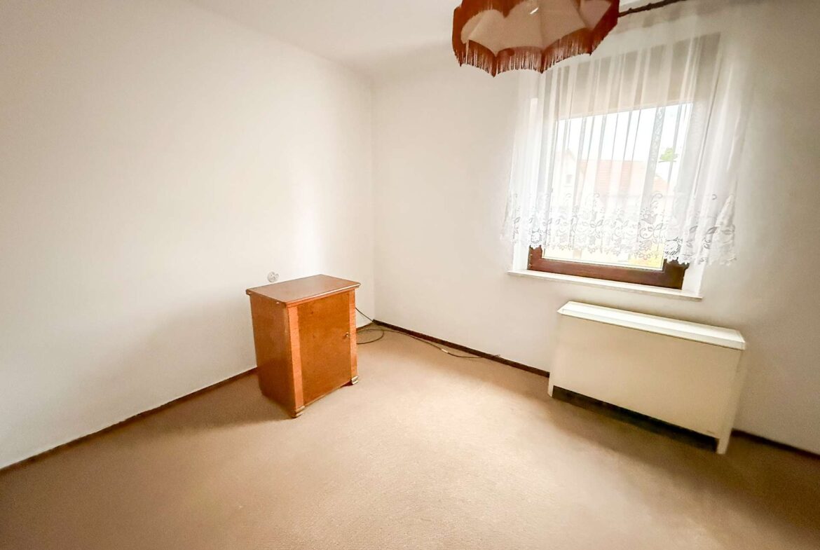 Empty beige room with a small wooden cabinet, window with lace curtains, and a wall heater beneath the window.”,