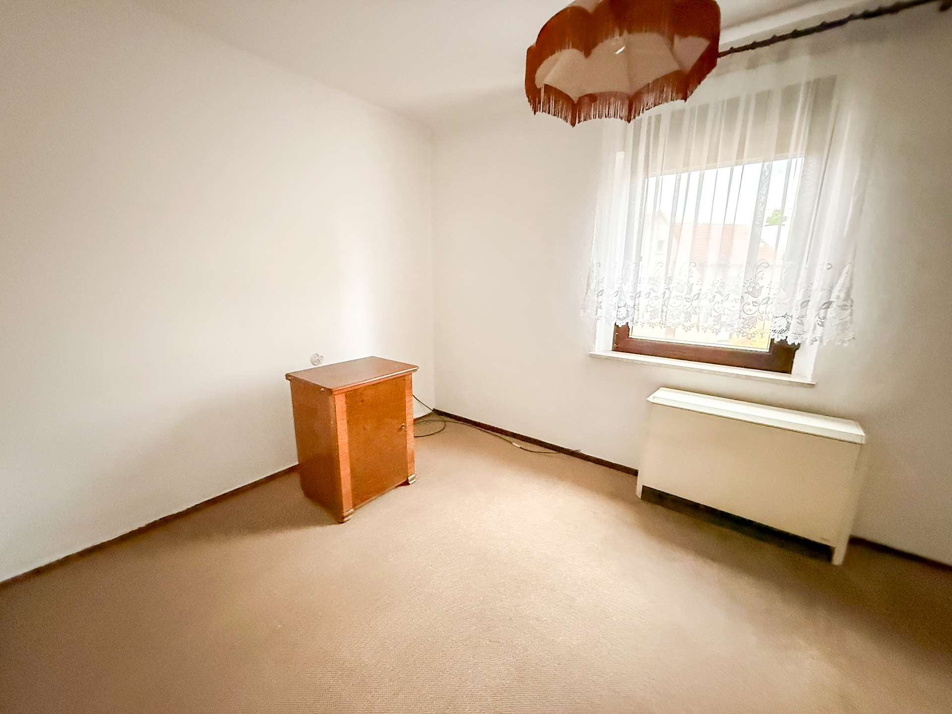 Empty beige room with a small wooden cabinet, window with lace curtains, and a wall heater beneath the window.”,