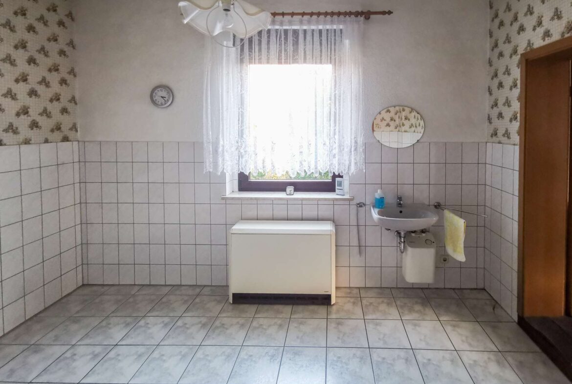 Bathroom with white tiled walls, lace-curtained window, round wall mirror, clock, and a small sink with a towel nearby.