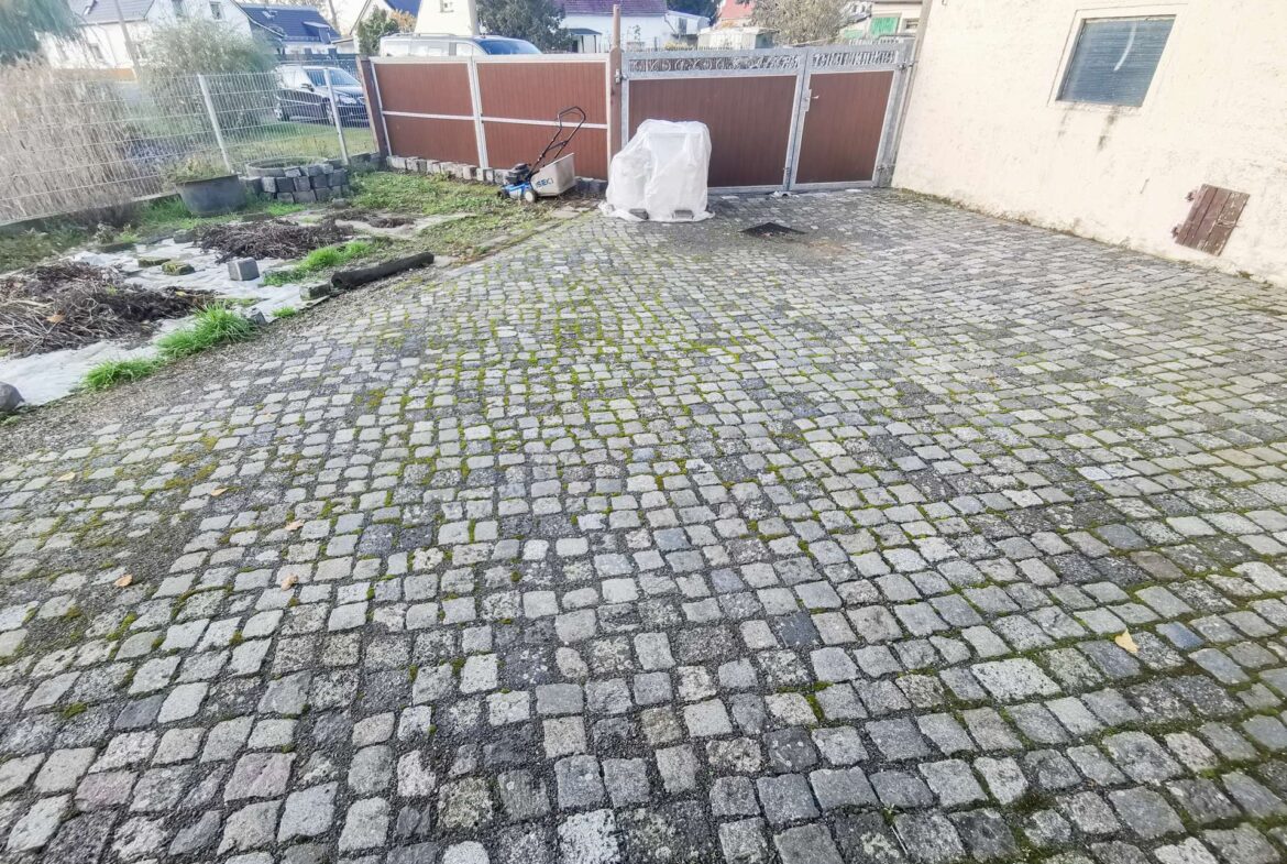 A cobblestone driveway with scattered debris, a small lawn mower, and a tarp-covered object near a brown gate; a pale building wall on the right.