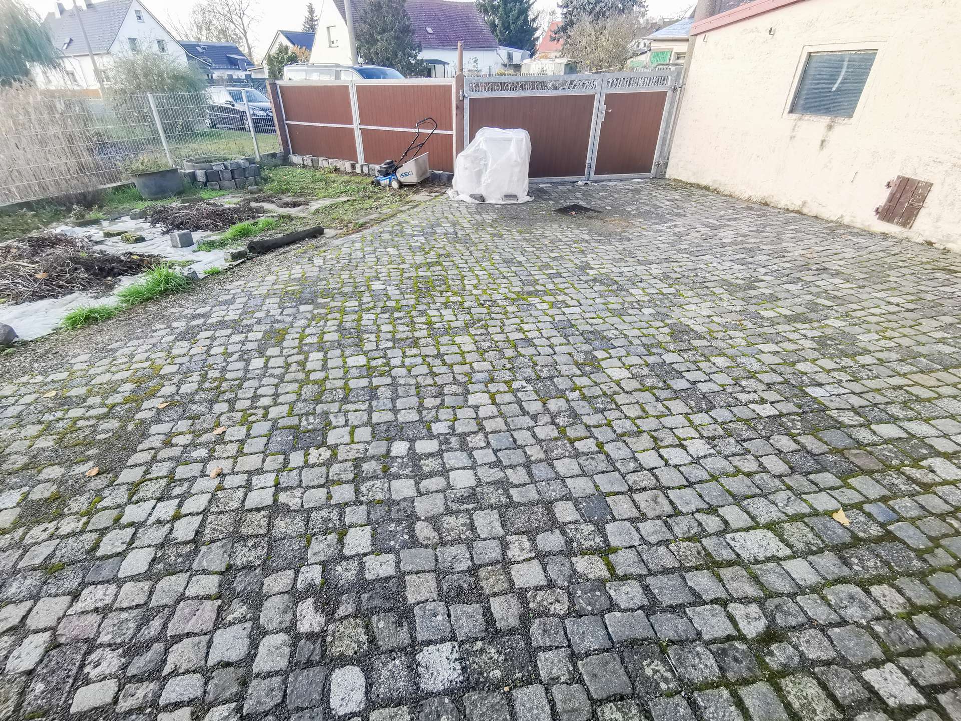 A cobblestone driveway with scattered debris, a small lawn mower, and a tarp-covered object near a brown gate; a pale building wall on the right.