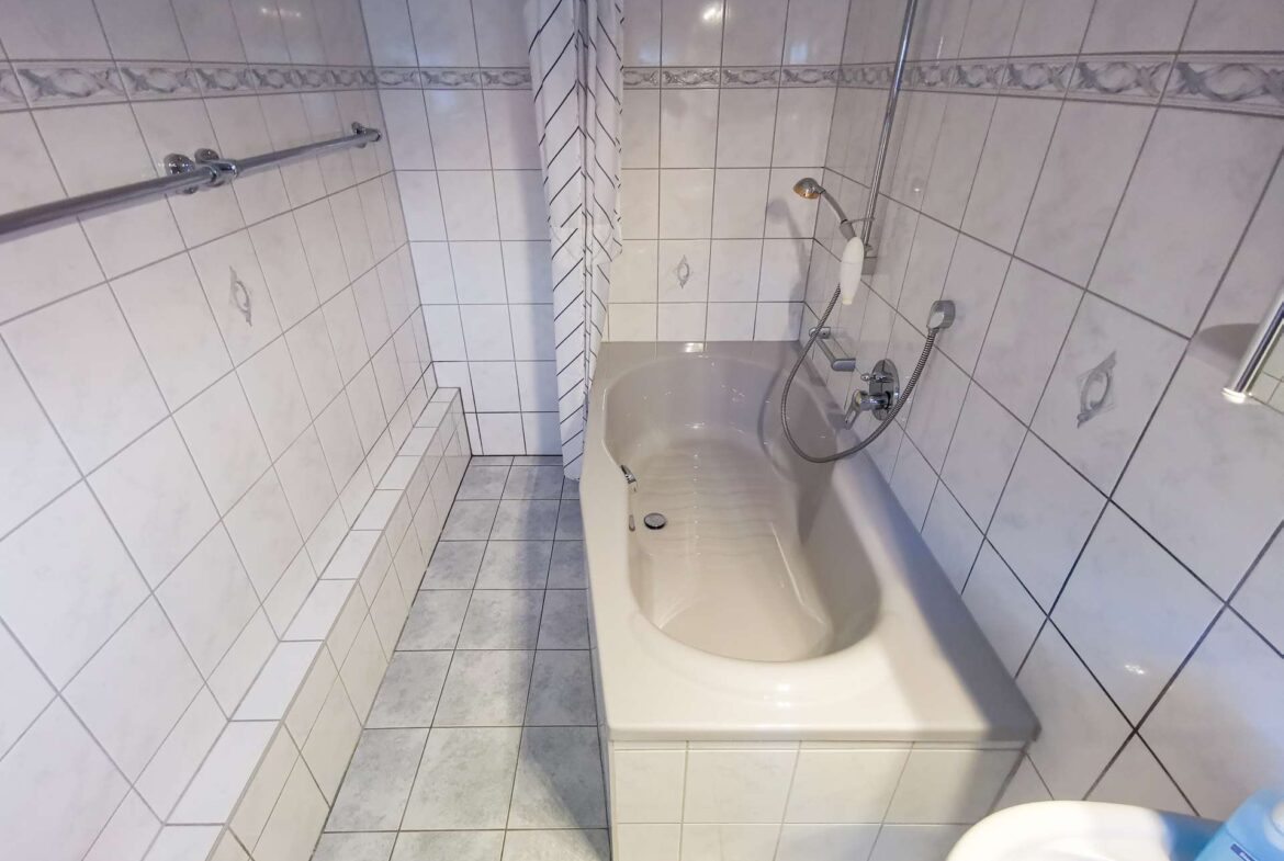 Narrow tiled bathroom with a built-in bathtub, handheld shower, striped shower curtain, and a chrome grab bar along the left wall.