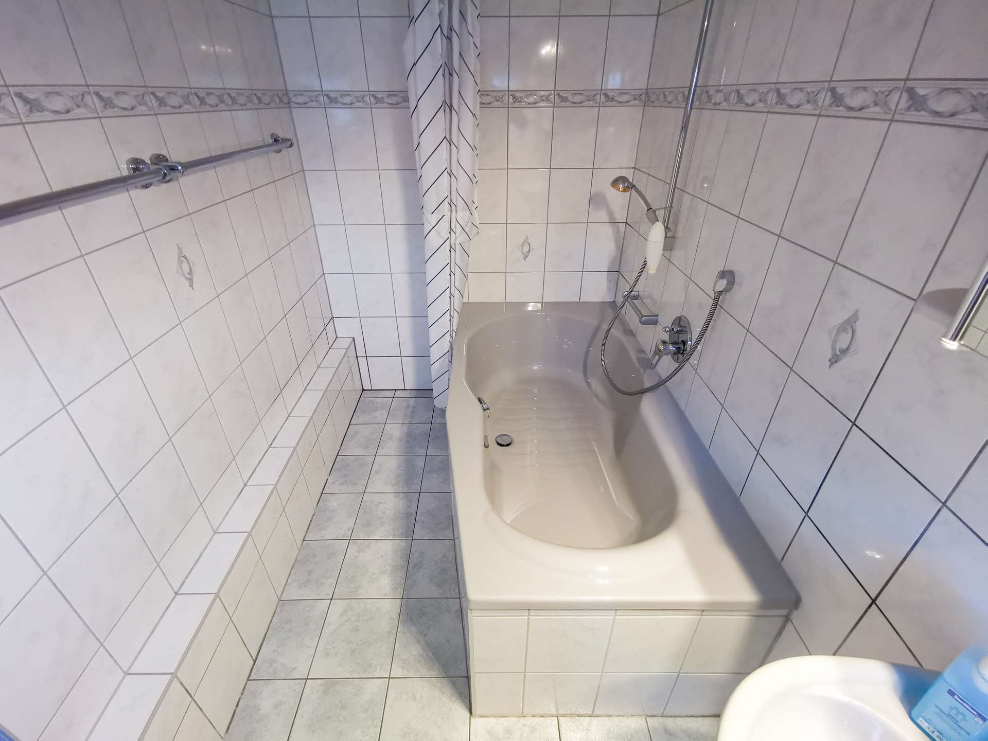 Narrow tiled bathroom with a built-in bathtub, handheld shower, striped shower curtain, and a chrome grab bar along the left wall.