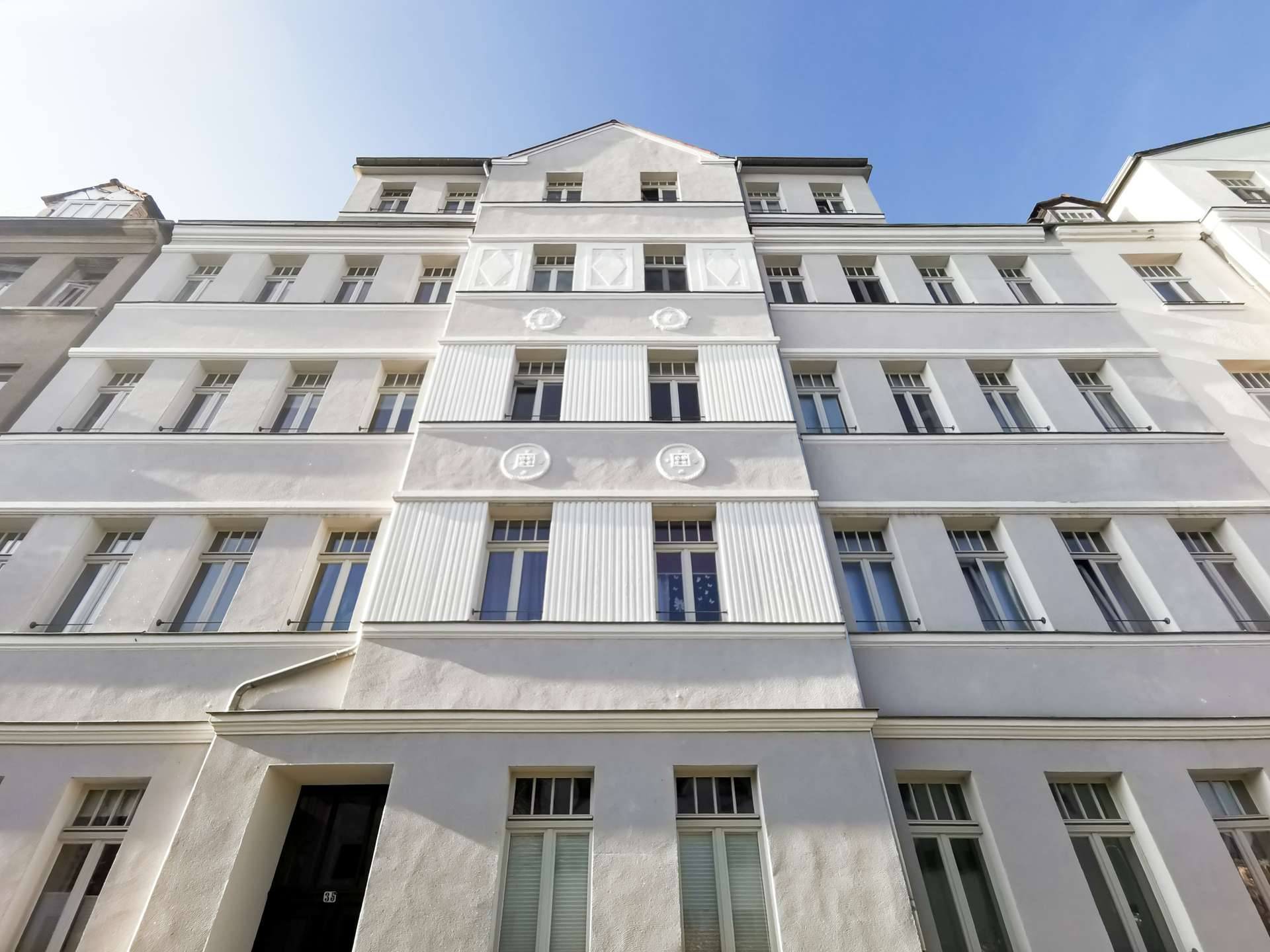 Upward-looking view of a white residential building with vertical fluted columns and decorative motifs on the façade against a clear blue sky.